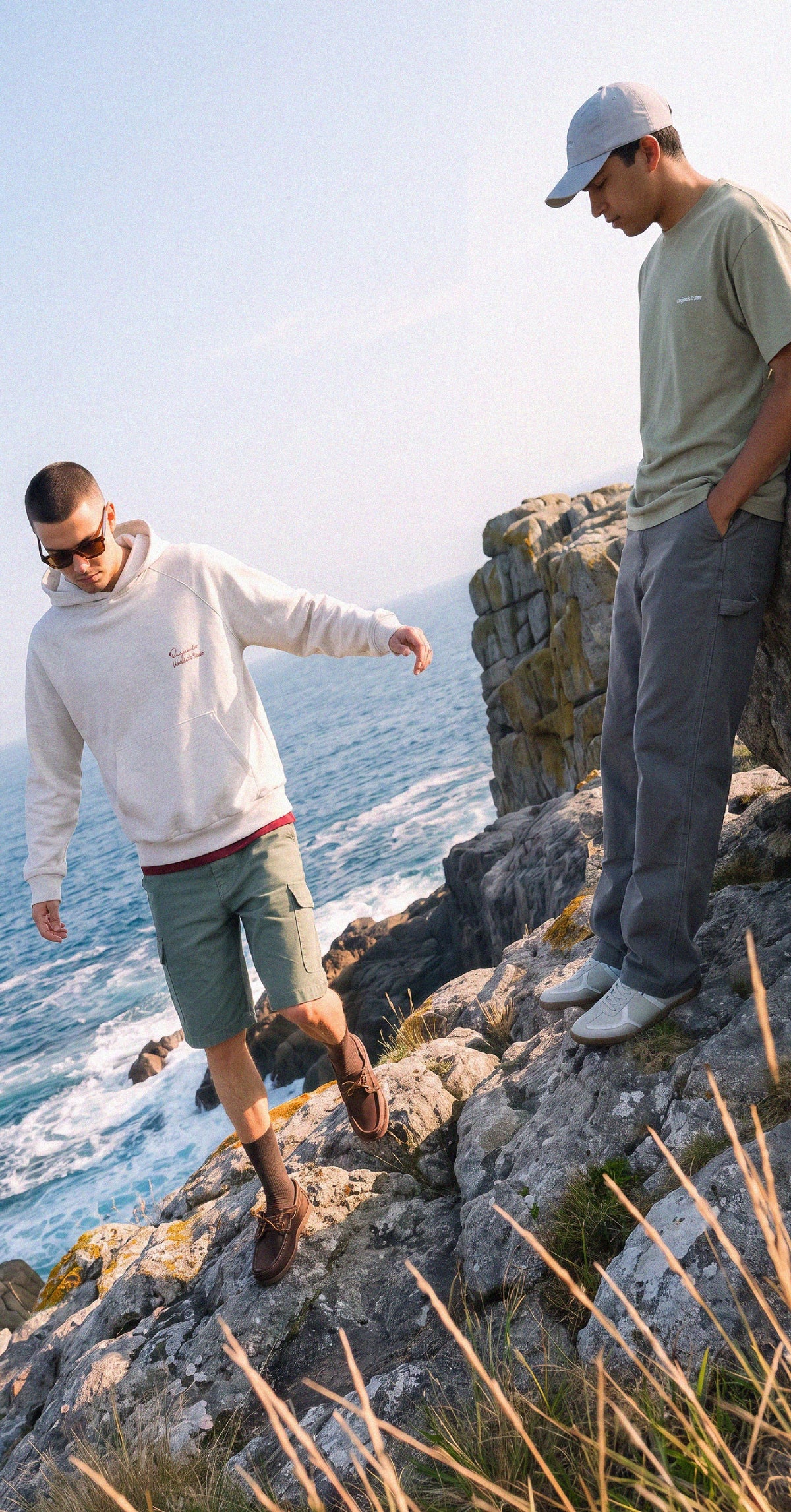 Dos modelos de Jack & Jones en la costa: buzo con capucha crudo y bermuda cargo verde; remera verde, gorra y pantalón gris